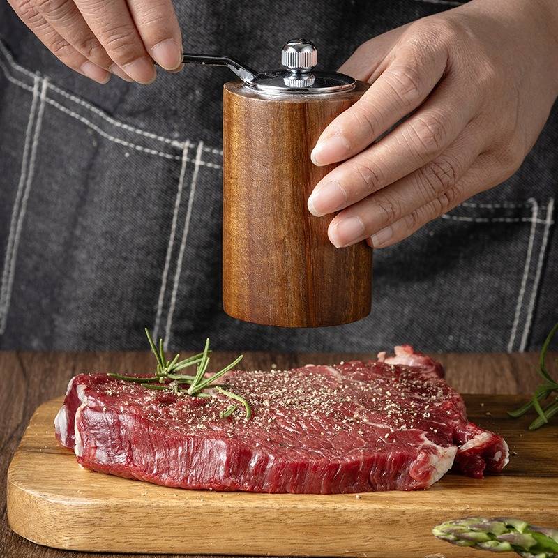 Hände würzen ein rohes Steak auf einem Holzschneidebrett mit einer Pfeffermühle, wobei Rosmarinzweige in der Nähe liegen.