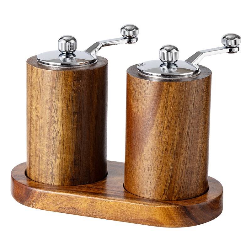 Zwei hölzerne manuelle Gewürzmühlen auf einer passenden Holzbasis, jede mit einer Metallkurbel und einem oberen Knopf.