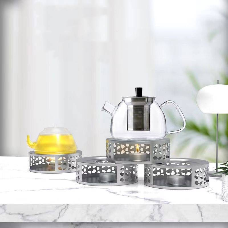 Glaskanne und Teetasse auf dekorativen Metallwärmesockeln auf einem weißen Marmortisch in einem hellen Raum präsentiert.