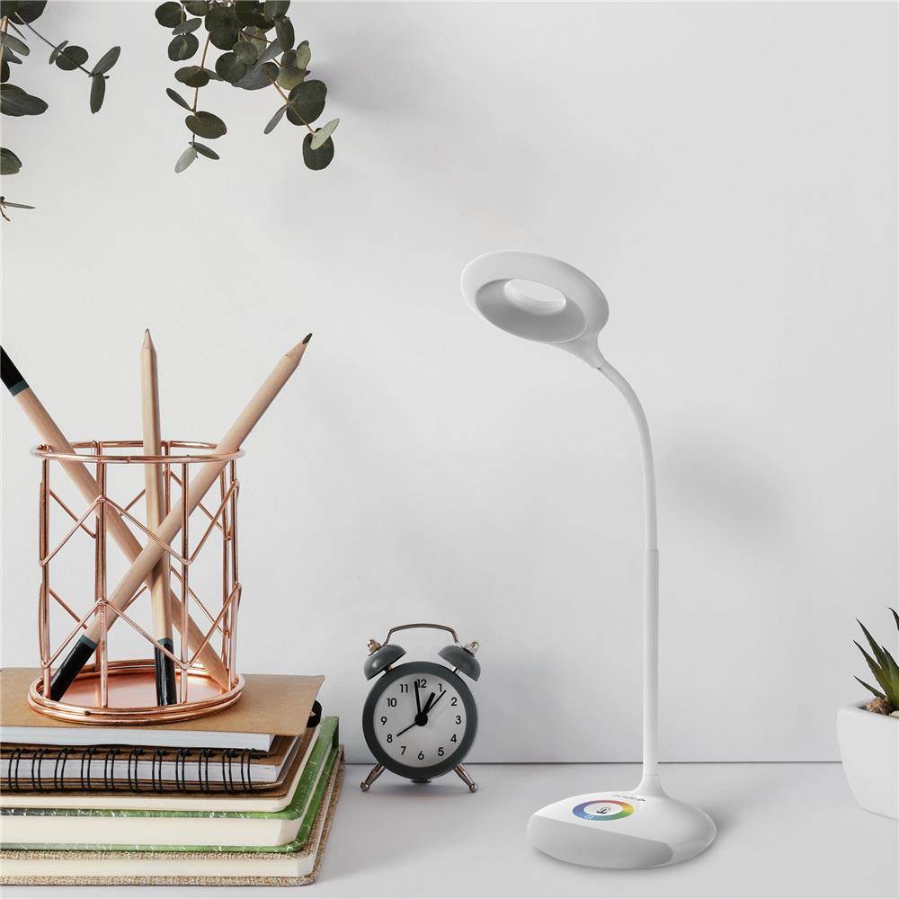 Minimalistischer Schreibtisch mit Stiftbecher auf gestapelten Notizbüchern, kleinem Wecker und weißer Schreibtischleuchte mit Ringen vor einer schlichten Wand.