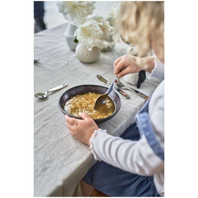 Ein Kind sitzt an einem Tisch und isst Suppe aus einer Schüssel mit einem Löffel.