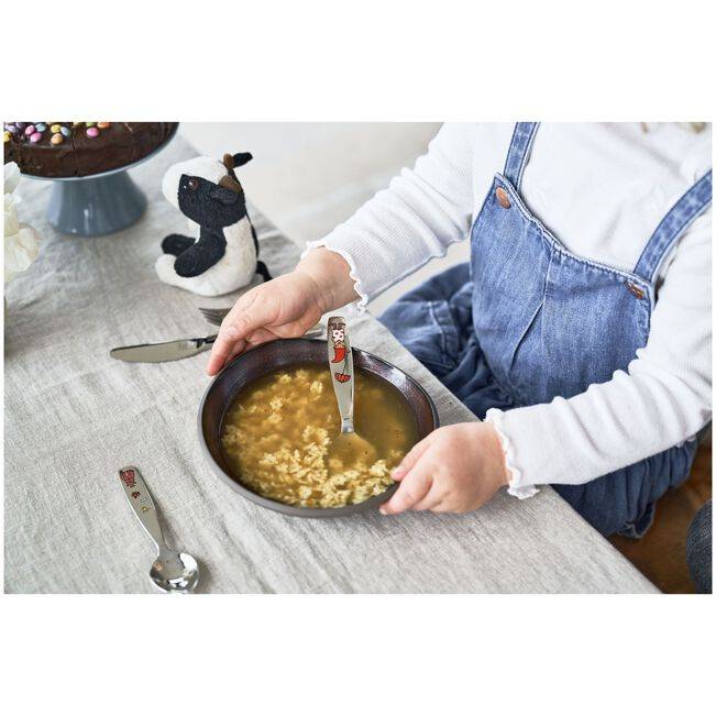 Kind in Jeans-Latzhose, das Suppe aus einer Schüssel an einem Tisch isst, mit einem Löffel und einer Servierschüssel in der Nähe.