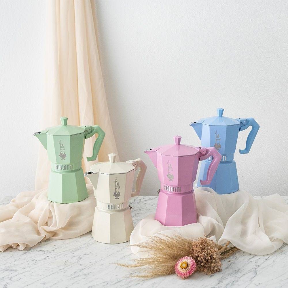 Vier farbenfrohe Moka-Kannen – grün, creme, rosa und blau – auf drapiertem Stoff mit getrockneten Blumen vor einer weißen Wand arrangiert.