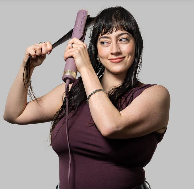Frau, die einen rosa Haarglätter verwendet, um einen Bereich ihres langen dunklen Haars vor einem einfachen grauen Hintergrund zu stylen.