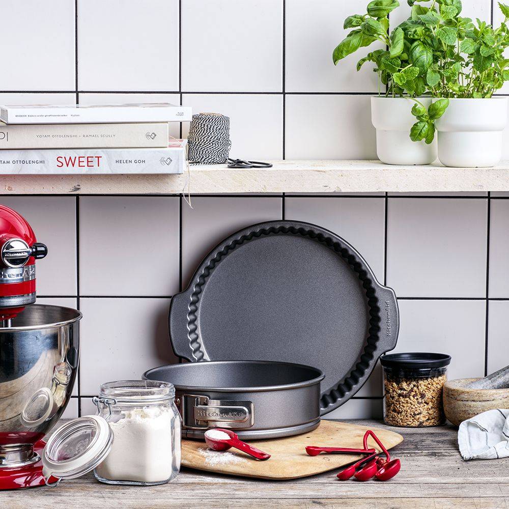 Küchenarbeitsplatte mit Backwerkzeugen und Zutaten: Küchenmaschine, Kuchenform, Tortenform, Gläser, Messlöffel und ein Topf mit Kräutern auf einem Regal.