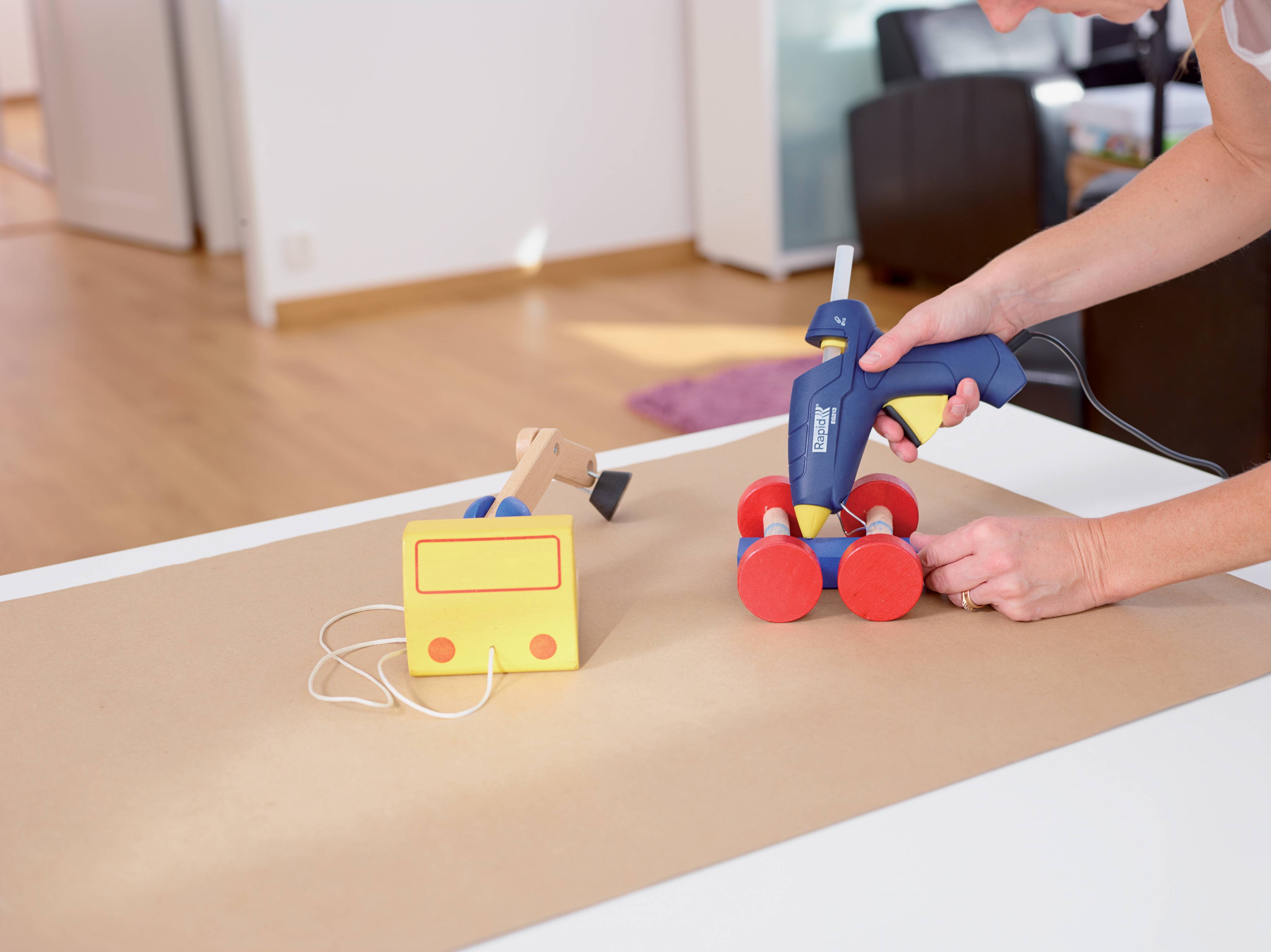 Hände, die eine Heißklebepistole verwenden, um ein kleines Spielzeugauto mit roten Rädern und einem gelben Batteriepack auf einer Werkbank zusammenzusetzen.
