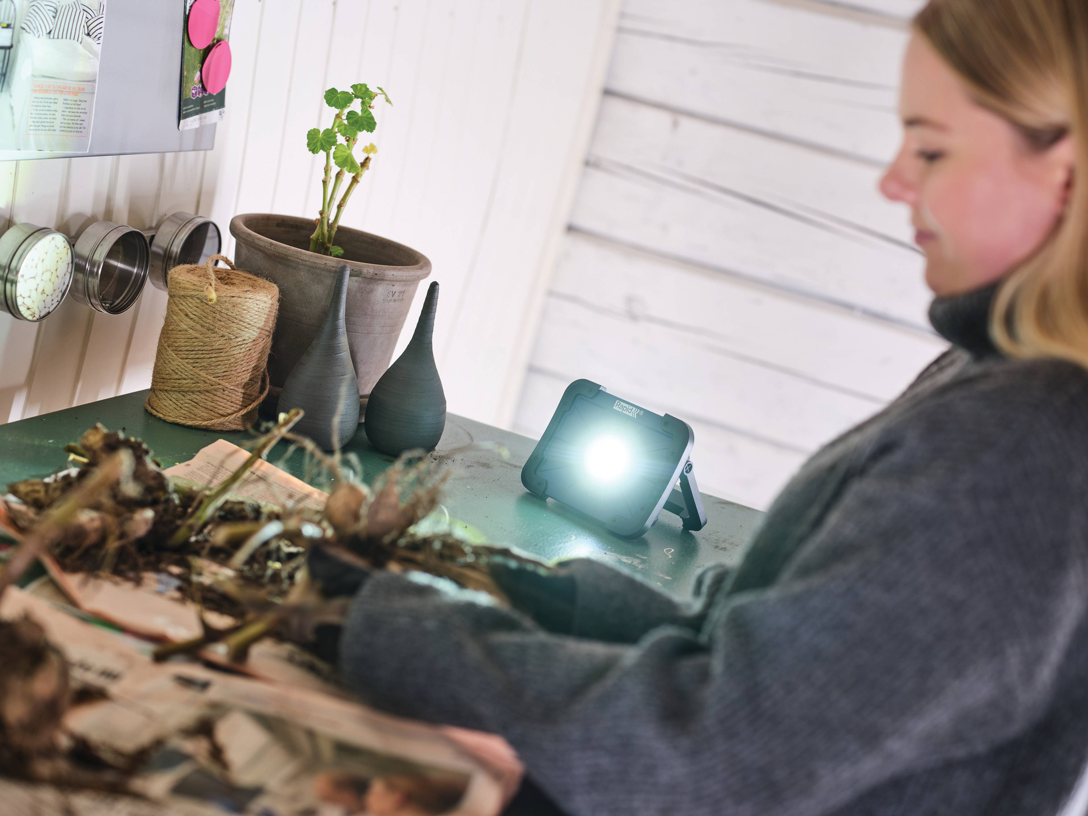 Eine Person untersucht eine Tischplattenausstellung aus getrockneten Blättern und Pflanzenmaterialien, beleuchtet von einer kleinen hellen Lampe in einer Werkstatt oder einem Studio.