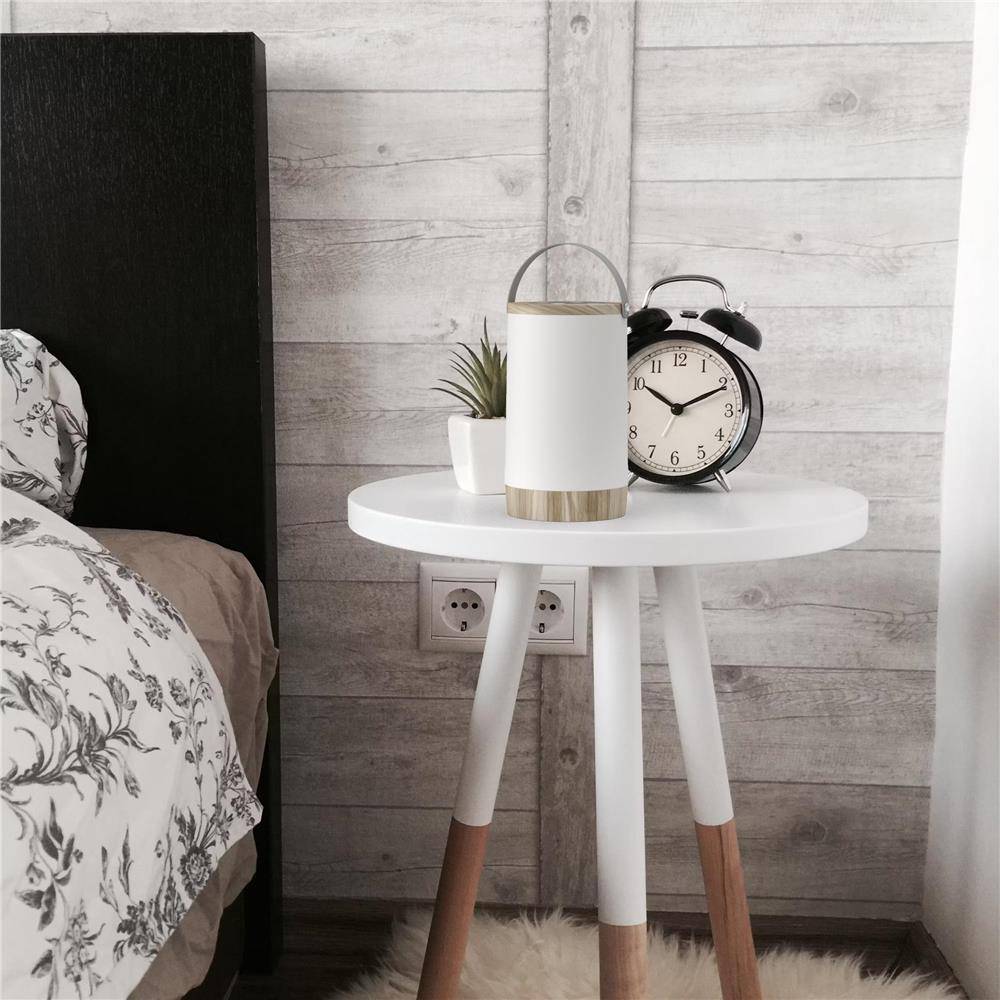Nachttisch mit einer Lampe, einer kleinen Pflanze und einem Wecker neben einem Bett vor einer holzgetäfelten Wand.