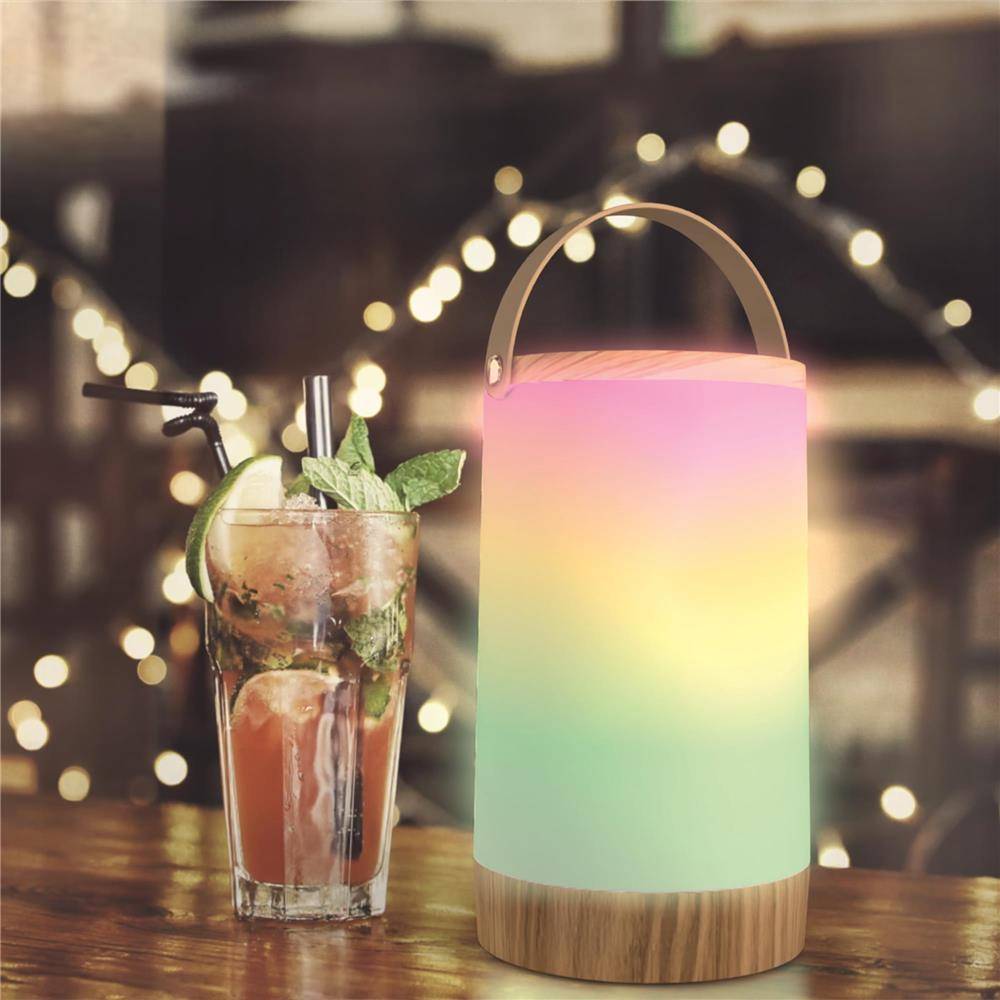 Ein Cocktail in einem hohen Glas mit Minze und Limette sitzt neben einer leuchtenden Regenbogen-Farbverlauf-Laterne auf einem Holztisch mit unscharfen Lichterketten im Hintergrund.