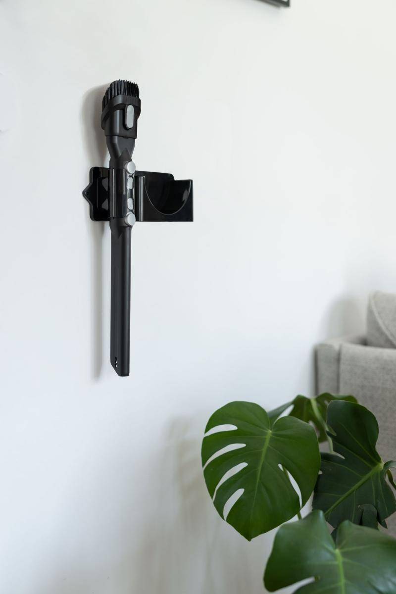 Schwarzer, an der Wand montierter Staubsaugerhalter an einer weißen Wand über einer grünen Monstera-Pflanze, mit einem Teil eines Sofas rechts.