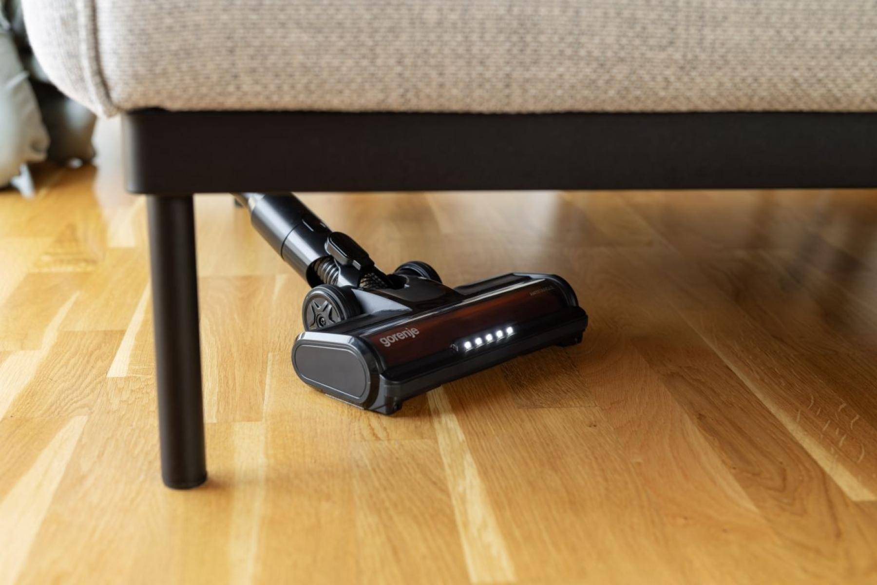 Schnurloser Staubsaugerkopf wird unter einem Sofa auf einem Hartholzboden gereinigt, wobei die niedrige Möbelfreiräumung demonstriert wird.
