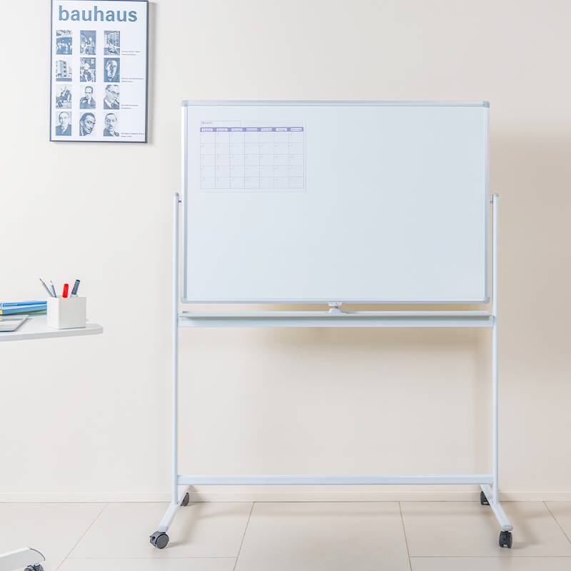 Leeres Klassenzimmer mit einem fahrbaren Whiteboard, auf dem ein kleines Kalenderraster zu sehen ist, neben einem „Bauhaus"-Plakat und einem kleinen Regal mit Schulbedarfsartikeln.