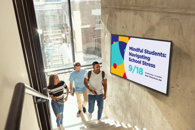Studenten gehen Treppen neben einem an der Wand montierten Bildschirm hinauf, auf dem am 18.9. „Mindful Students: Navigating School Stress" (Achtsamkeitsvolle Studenten: Umgang mit Schulstress) beworben wird.