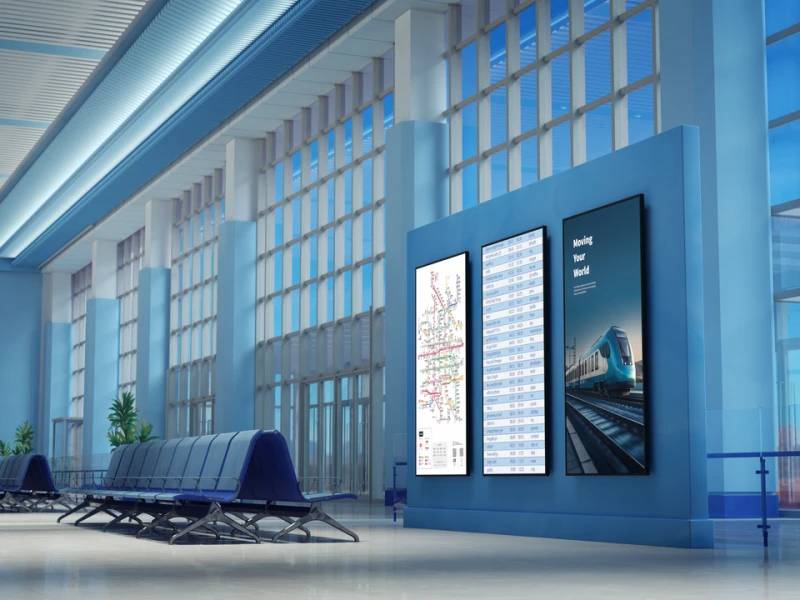 Moderner Bahnhofskomplex mit Sitzreihen und drei Wandplakaten, darunter eine Verkehrskarte, eine Fahrplantafel und eine Zugwerbung.