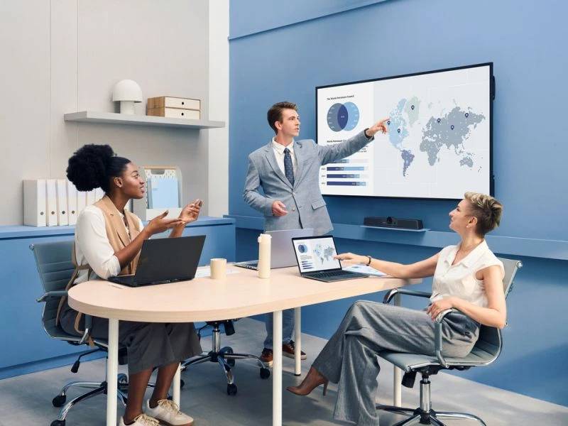 Geschäftsteam in einem Konferenzraum, das ein Weltkarten-Daten-Dashboard auf einem Wandbildschirm überprüft, wobei ein Mann präsentiert und zwei Frauen an einem Tisch mit Laptops sitzen.