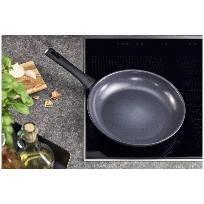 Antihaft-Bratpfanne auf einer Kochplatte neben Knoblauch, Kräutern und Öl auf einer Küchentheke.
