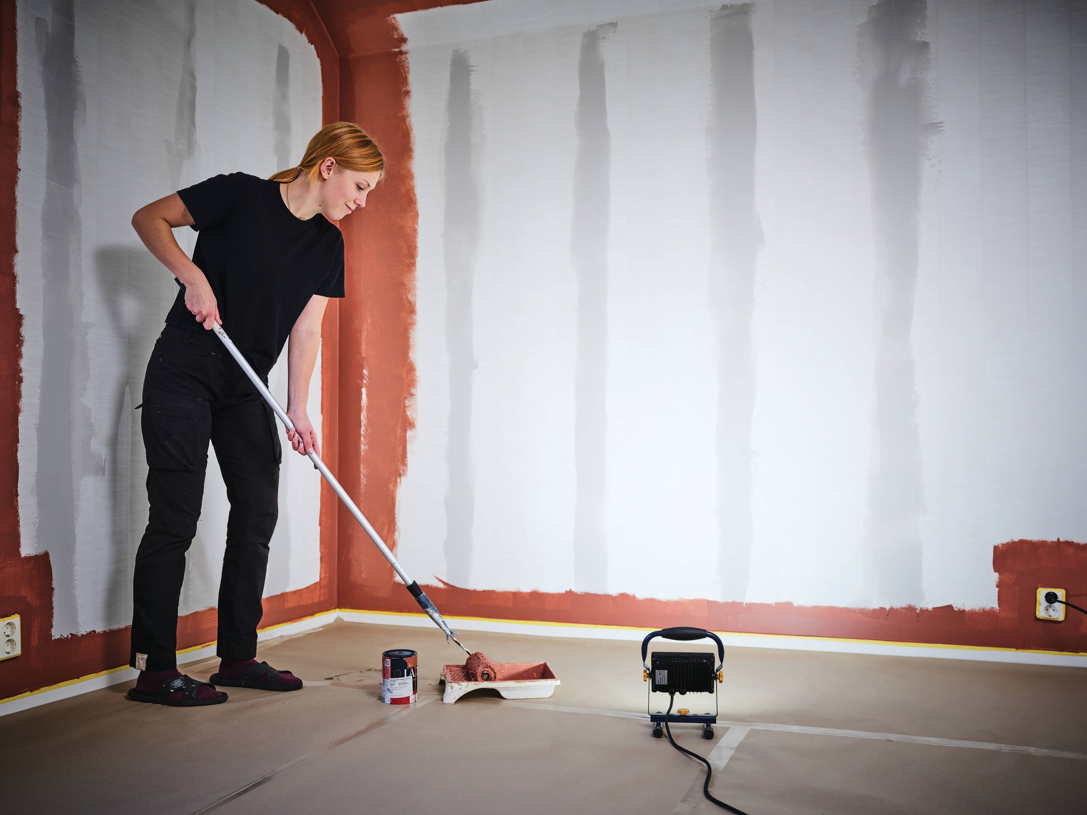 Eine Person streicht eine Zimmerwand mit einer Farbwalze und trägt rote Grundierung über weiße Gipskartonplatten in einem unfertigen Innenraum auf.