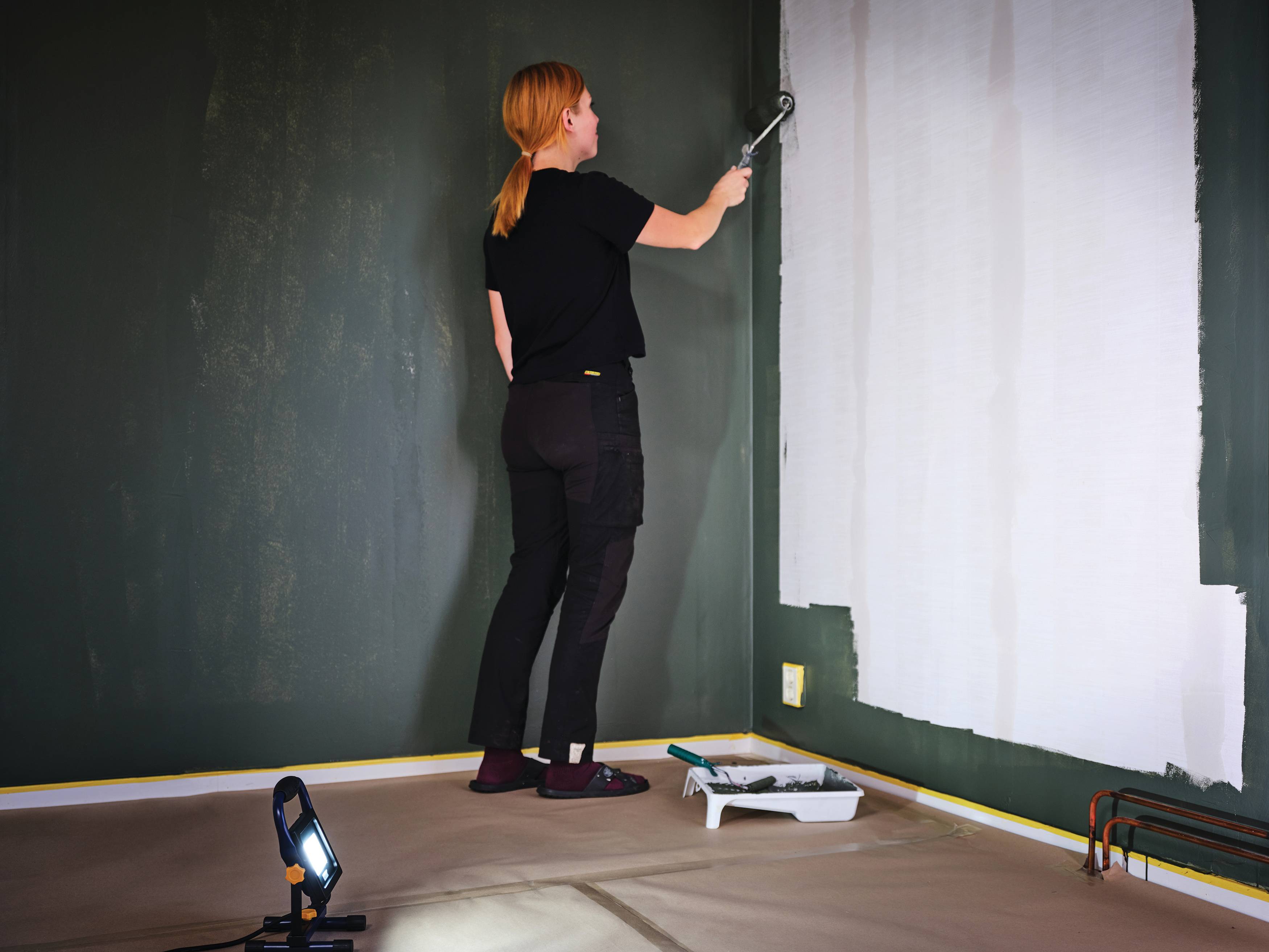 Eine Person malt eine Wand mit einer Rolle an und bedeckt eine dunkelgrüne Raumwand mit weißer Farbe, während sie auf einem geschützten Boden steht.