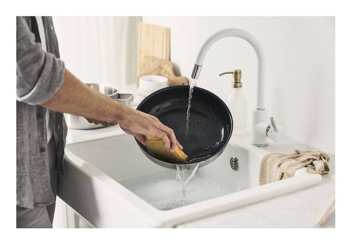 Eine Person spült eine schwarze Bratpfanne in einer Küchenspüle unter fließendem Wasser, während sie einen Schwamm hält und Geschirr wäscht.