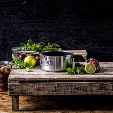 Eine Edelstahlkasserolle sitzt auf einem rustikalen Holztisch mit einer Limette, Kräutern und einem Glasgefäß, was auf Kochzutaten oder Lebensmittelzubereitung hindeutet.