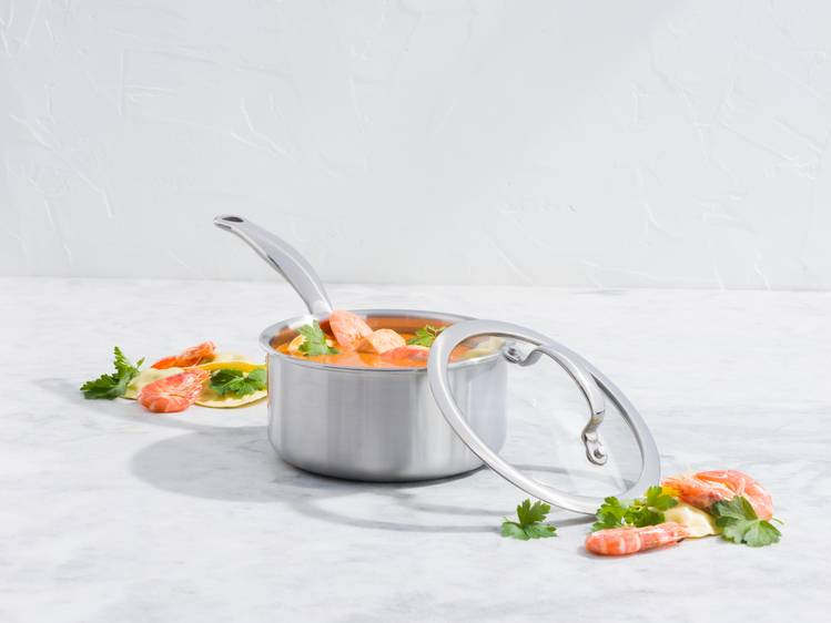 Ein Metalltopf mit Garnelen- und Gemüsesuppe mit geöffnetem Deckel, garniert mit Petersilie auf einer weißen Oberfläche.
