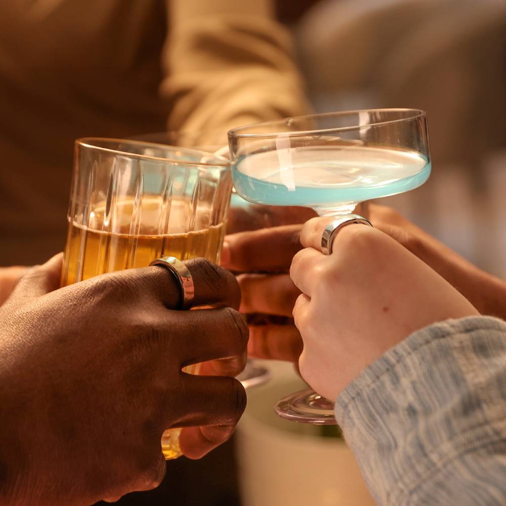 Drei Hände mit Ringen stoßen mit bunten Cocktailgläsern in warmem Licht an.