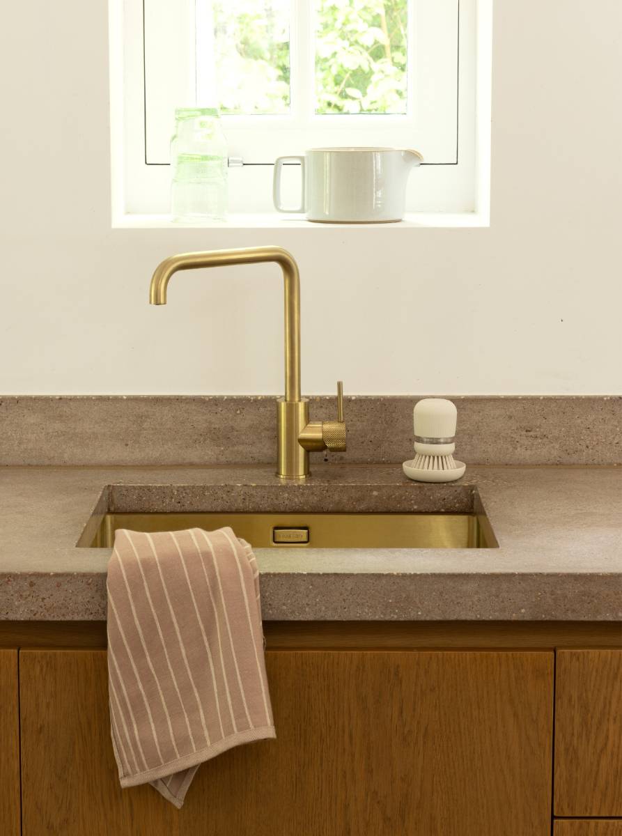 Modernes Spülbecken mit Messingarmaturen, gestreiftes Handtuch über dem Becken drapiert, Scheuerbürste daneben und ein Topf auf der Fensterbank darüber.