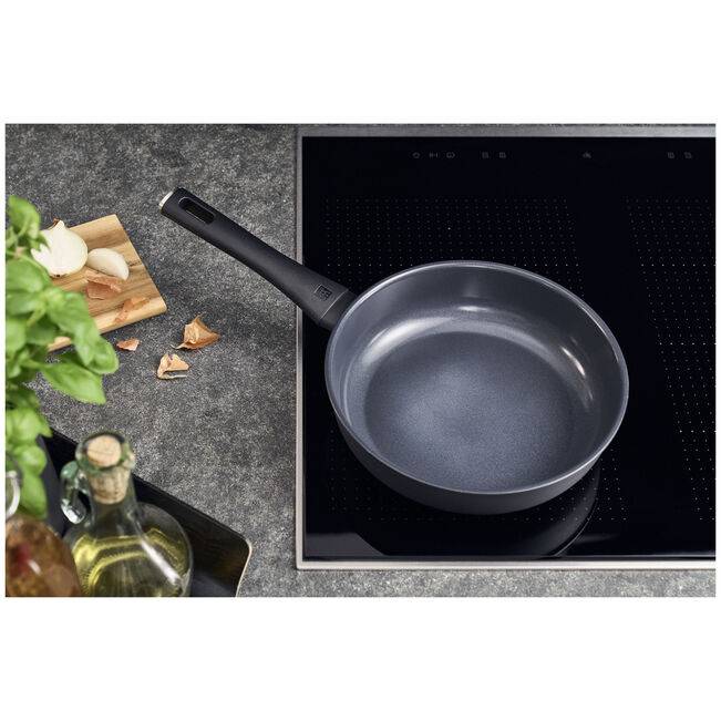 Leere antihaftbeschichtete Bratpfanne auf einem schwarzen Induktionskochfeld, mit Knoblauch, Kräutern und Ölflaschen auf der Arbeitsplatte daneben.