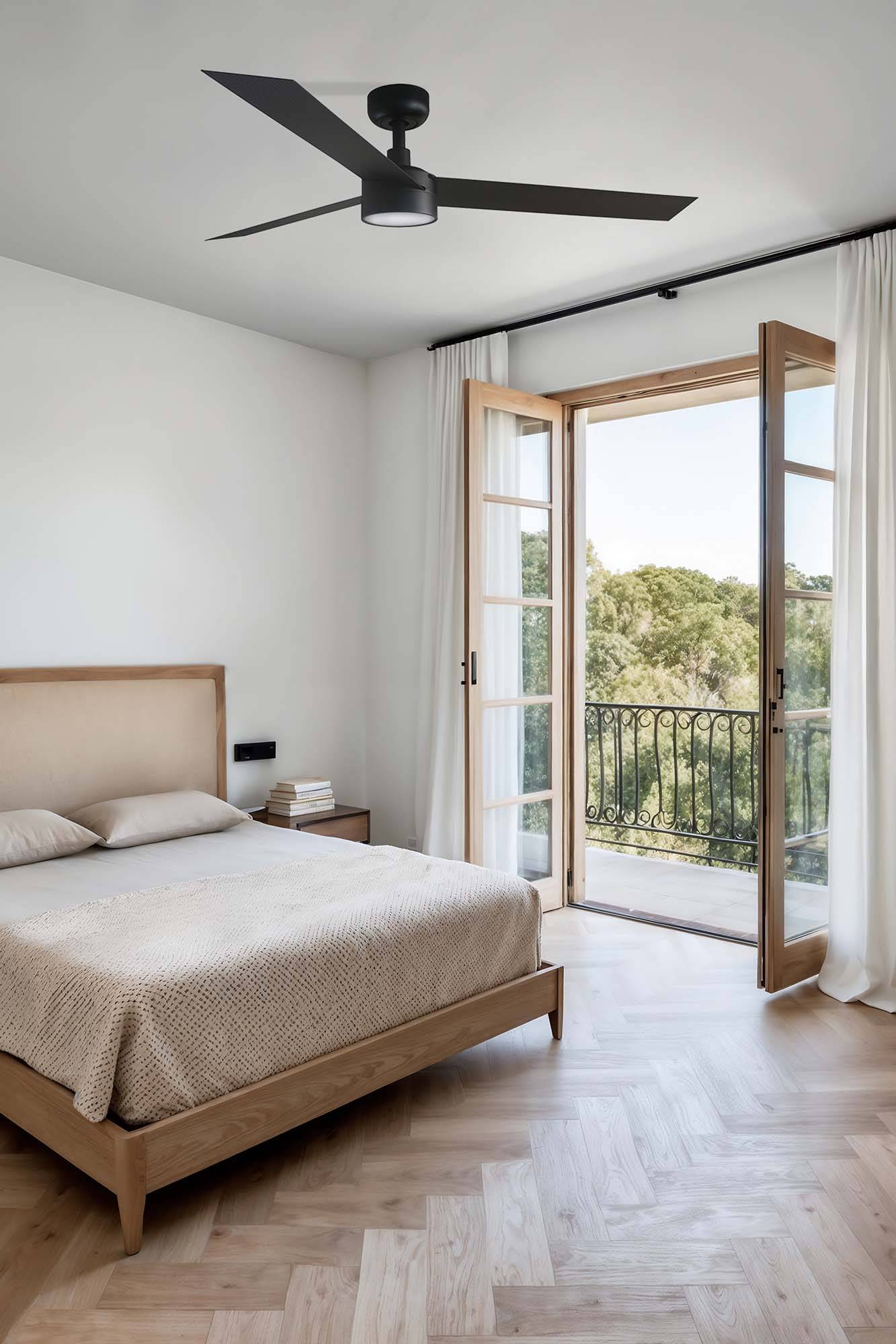 Minimalistisches Schlafzimmer mit einem Bett, Nachttisch und Büchern, schwarzem Deckenventilator und offenen französischen Türen, die zu einem Balkon mit Baumkronenblick führen.