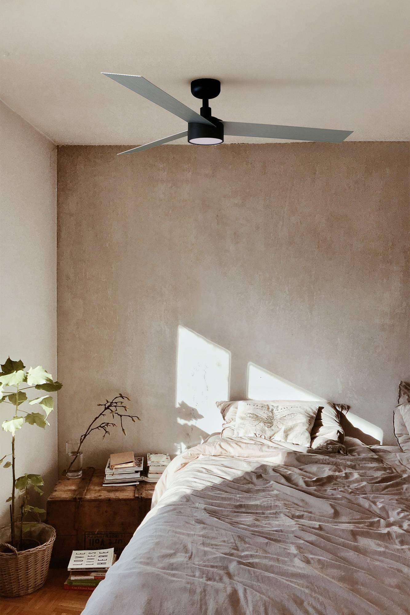 Ein minimalistisches Schlafzimmer mit einem ungemachten Bett, beiger strukturierter Wand, modernem Deckenventilator und Sonnenlicht, das einen geometrischen Schatten neben Bettnachttisch-Büchern und Pflanzen wirft.