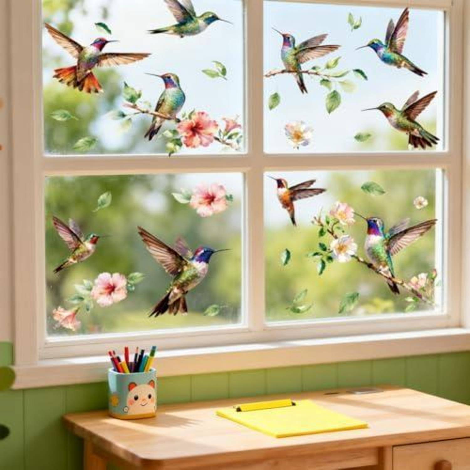 Fenster dekoriert mit bunten Kolibris und Blumen, über einem Kinderschreibtisch mit einem Stiftbecher und gelbem Notizbuch.