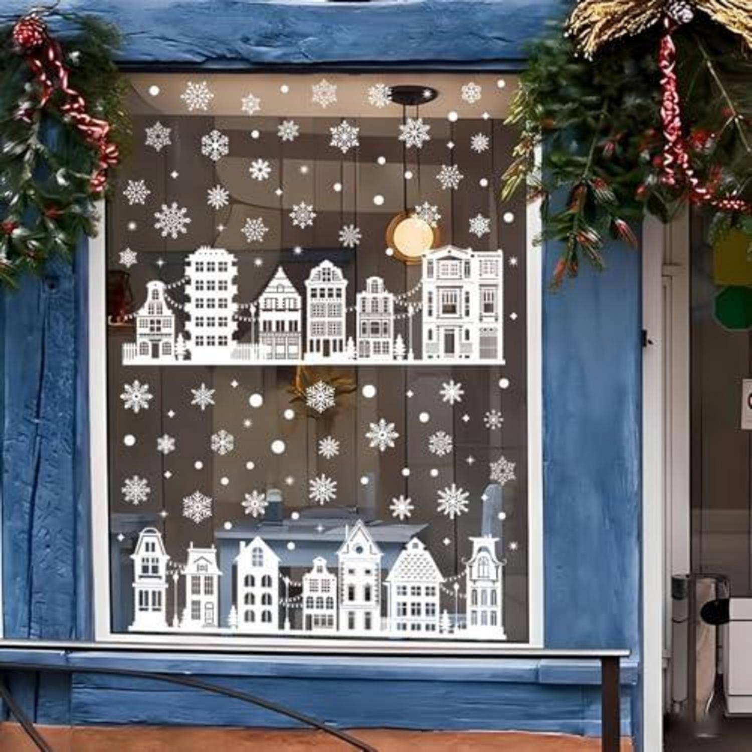 Mit Schneeflöckchen bedecktes Schaufenster, das mit weißen Papierhausausschnitten und festlichem Grün dekoriert ist und eine Winterdorfszene schafft.