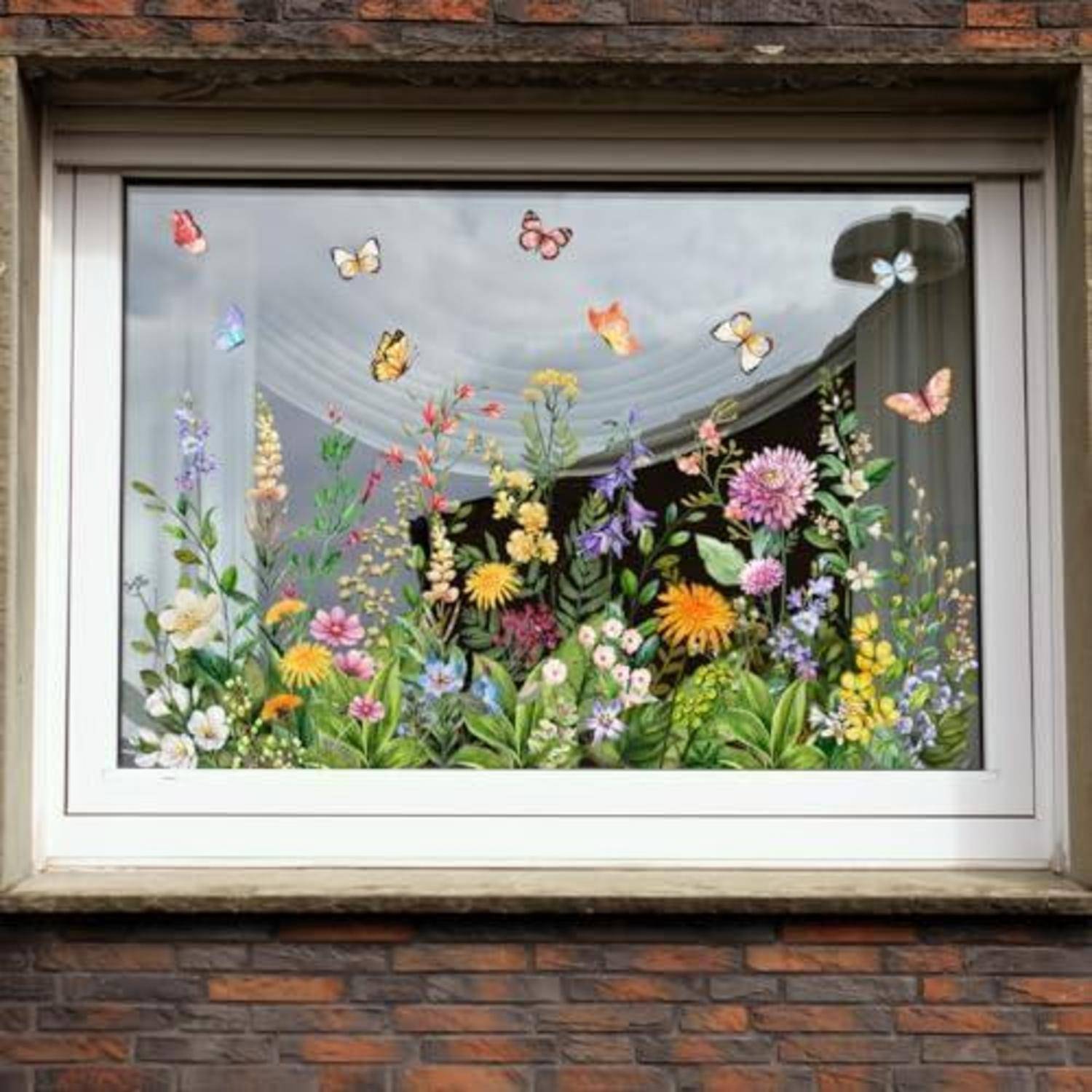 Fensterausstellung mit farbenfrohen bemalten Blumen und Schmetterlingen in der unteren Hälfte, eingebettet in ein weißgerahmtes Fenster in einer Backsteinmauer.