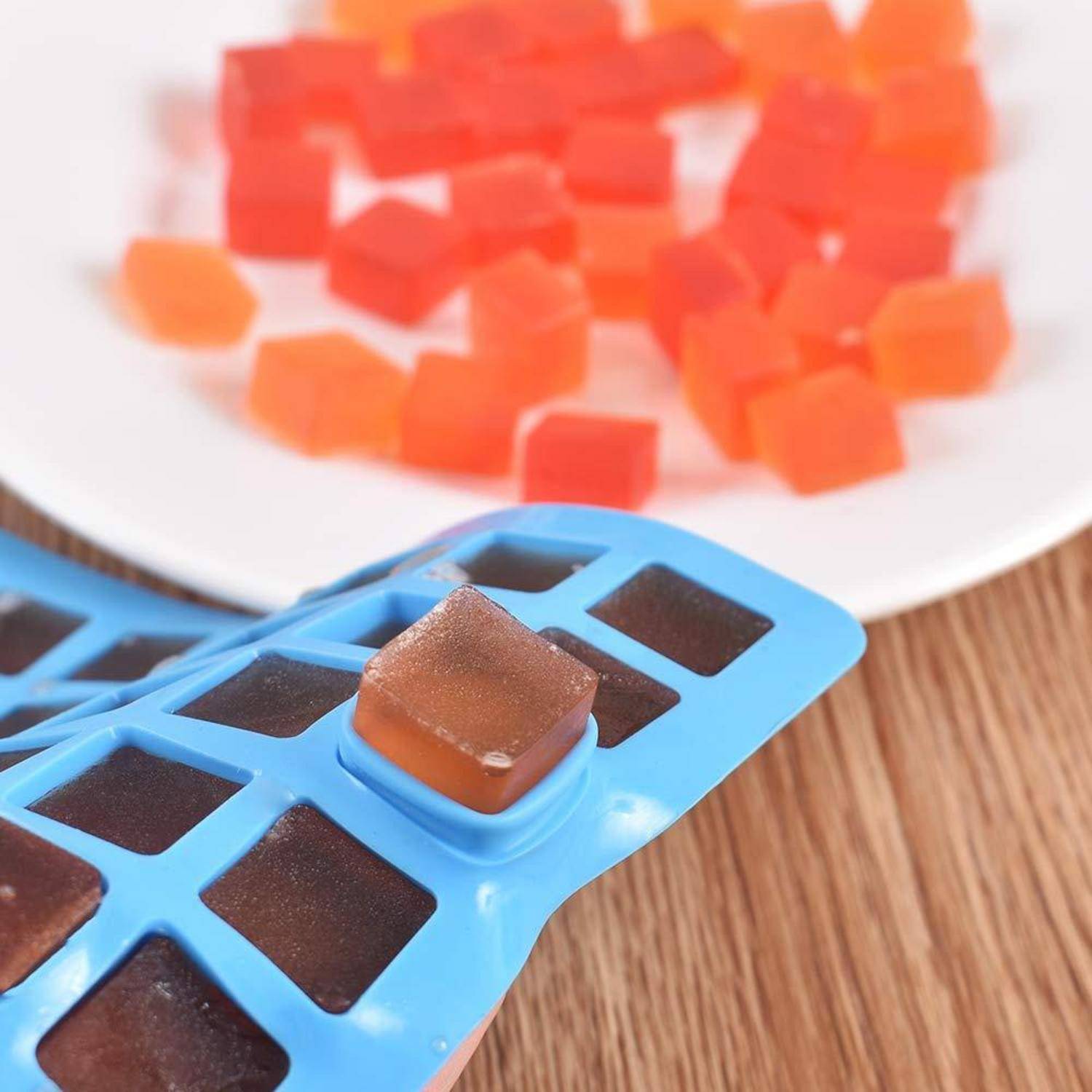 Blaues Silikon-Eiswürfeltablett mit einem braunen Würfel, der gerade herausgedrückt wird, und orangefarbenen Würfeln auf einem Teller im Hintergrund.