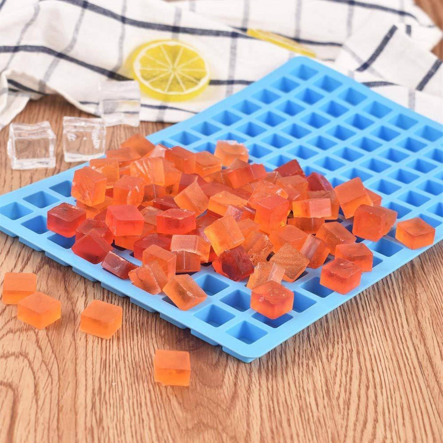 Orange durchsichtige Würfel auf einem blauen Silikon-Eiswürfelbehälter, mit einem Tuch mit Zitronendruck im Hintergrund und einigen Würfeln, die auf einem Holztisch verstreut sind.
