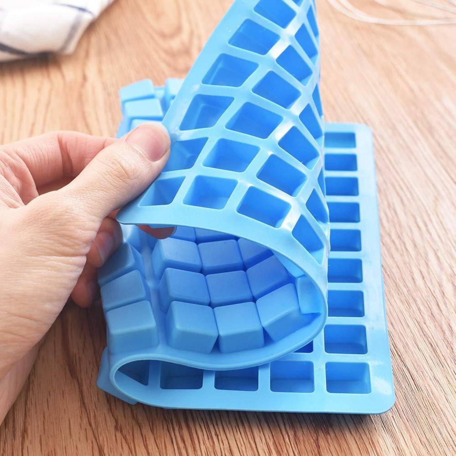 Hand, die eine flexible blaue Silikon-Eiswürfelform auf einer Holzoberfläche zurückzieht und einzelne Würfelfächer offenbart.