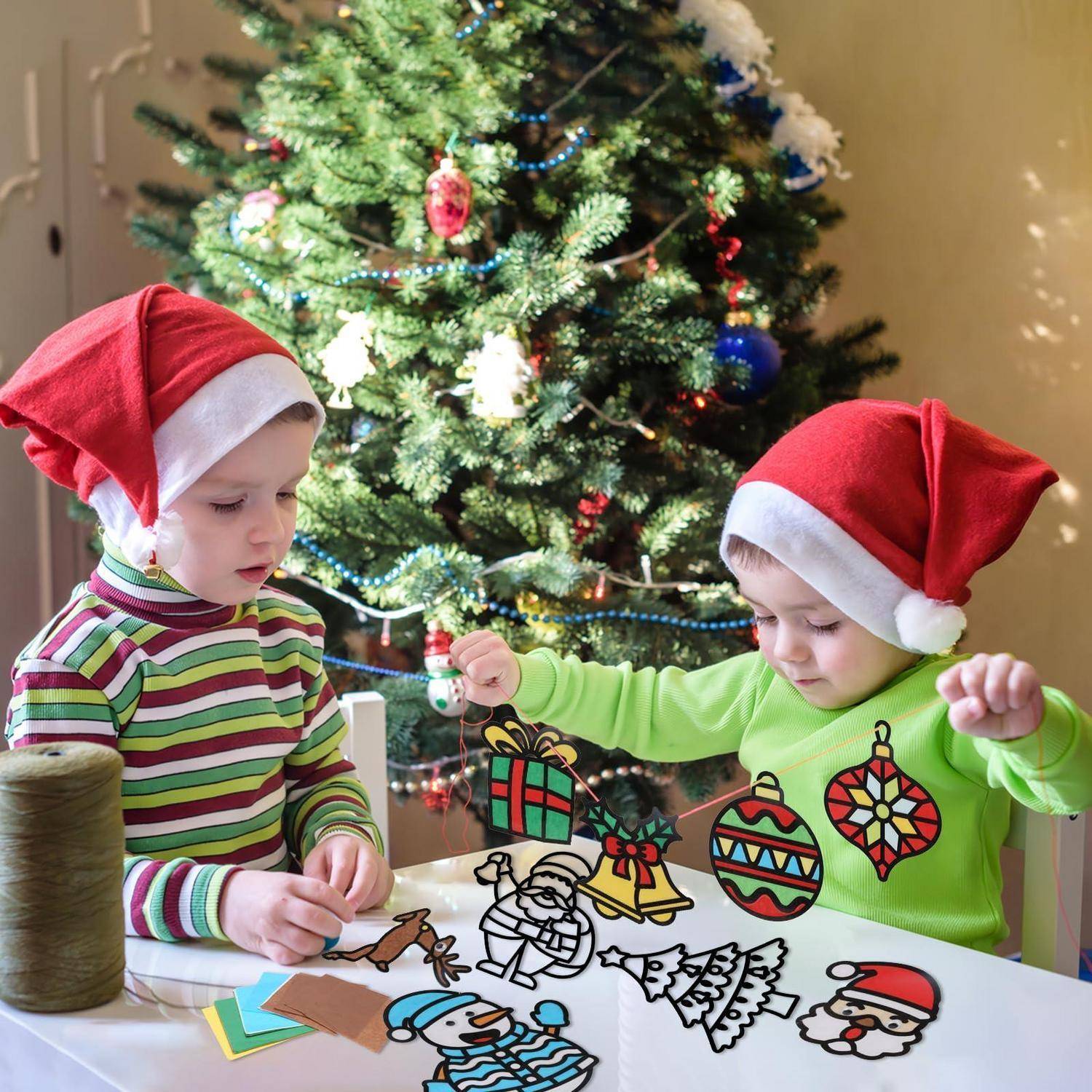 Zwei Kleinkinder in Weihnachtsmützen dekorieren eine Weihnachtsbaumbastelszene und platzieren bunte Weihnachtsausschnitte auf einem Tisch.