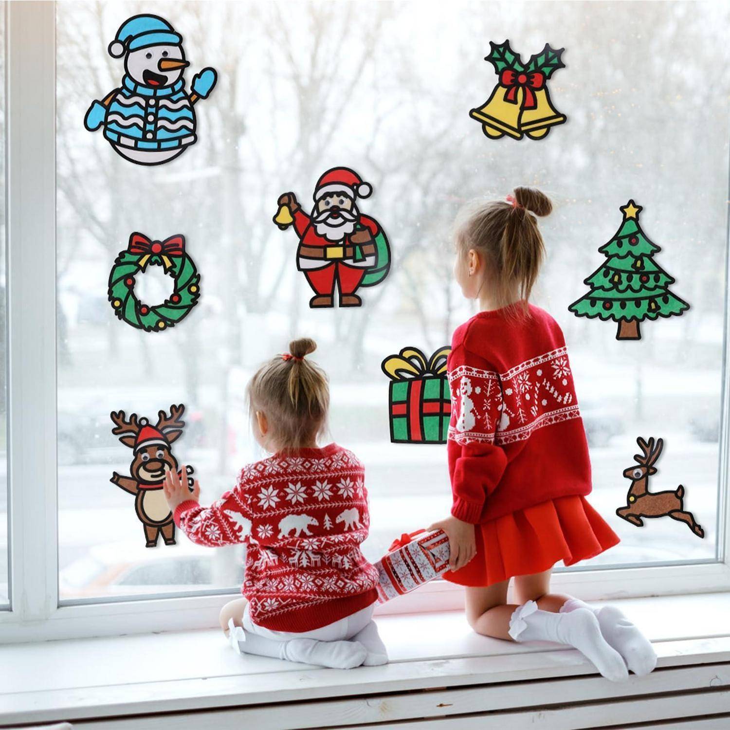 Zwei Kinder in roten Feiertagspullovern dekorieren ein Fenster mit Weihnachtsstickern, darunter der Weihnachtsmann, ein Kranz, Glocken, ein Baum, ein Geschenk und Rentiere.