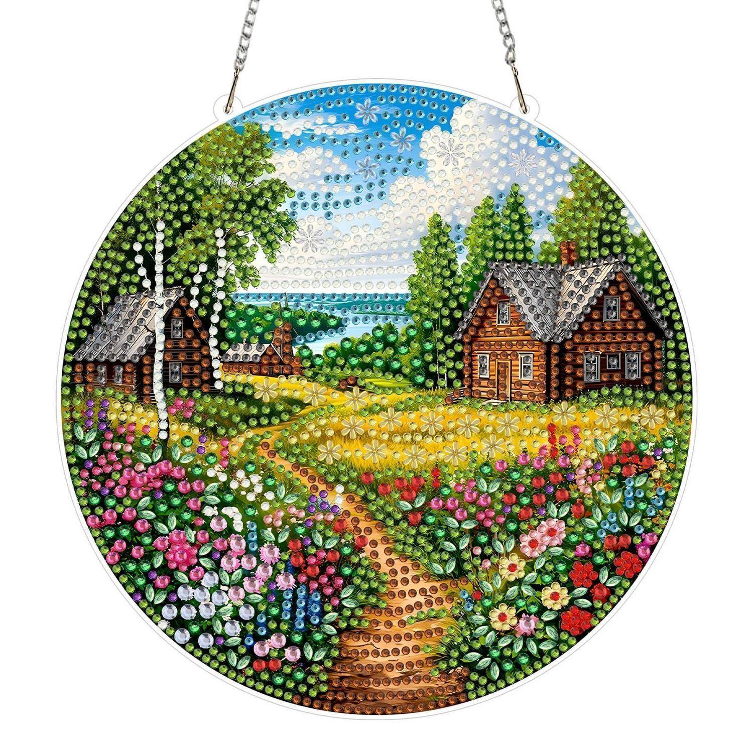 Rundes Hängdekorationskunstwerk, das eine ländliche Landschaft mit zwei Hütten, einem Feldweg, Blumen, Bäumen und einem See unter blauem Himmel zeigt.