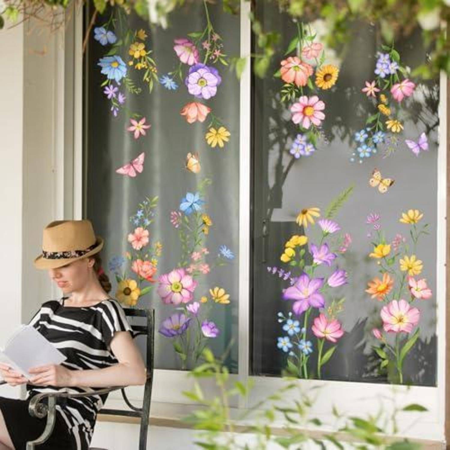 Eine Frau in einem Strohhut liest Papiere, während sie auf einem Veranda-Stuhl neben Fenstern sitzt, die mit bunten Papierblumen und Schmetterlingen geschmückt sind.