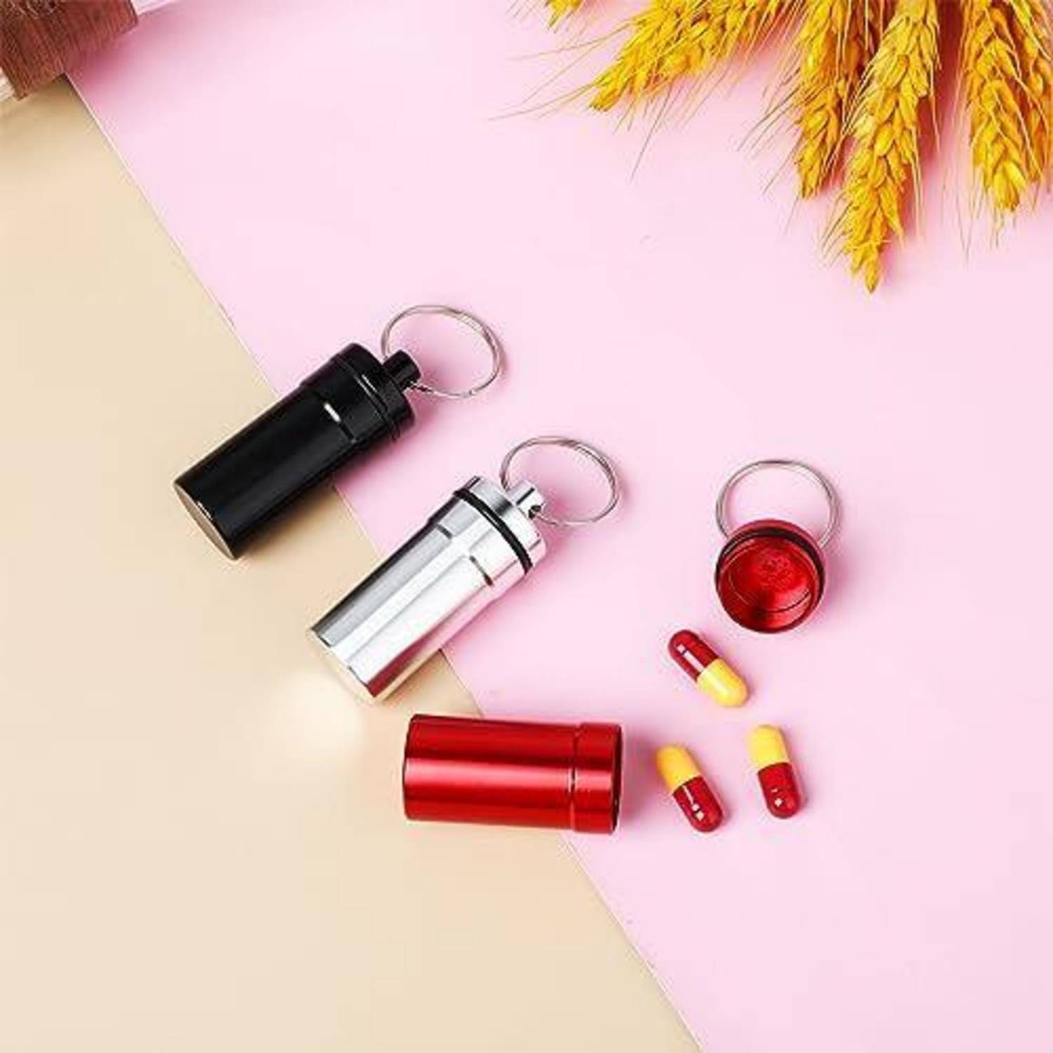 Drei Schlüsselanhänger-Pillendosen in Schwarz, Silber und Rot mit losen rot-gelben Kapseln auf einem rosa und beigen Hintergrund.