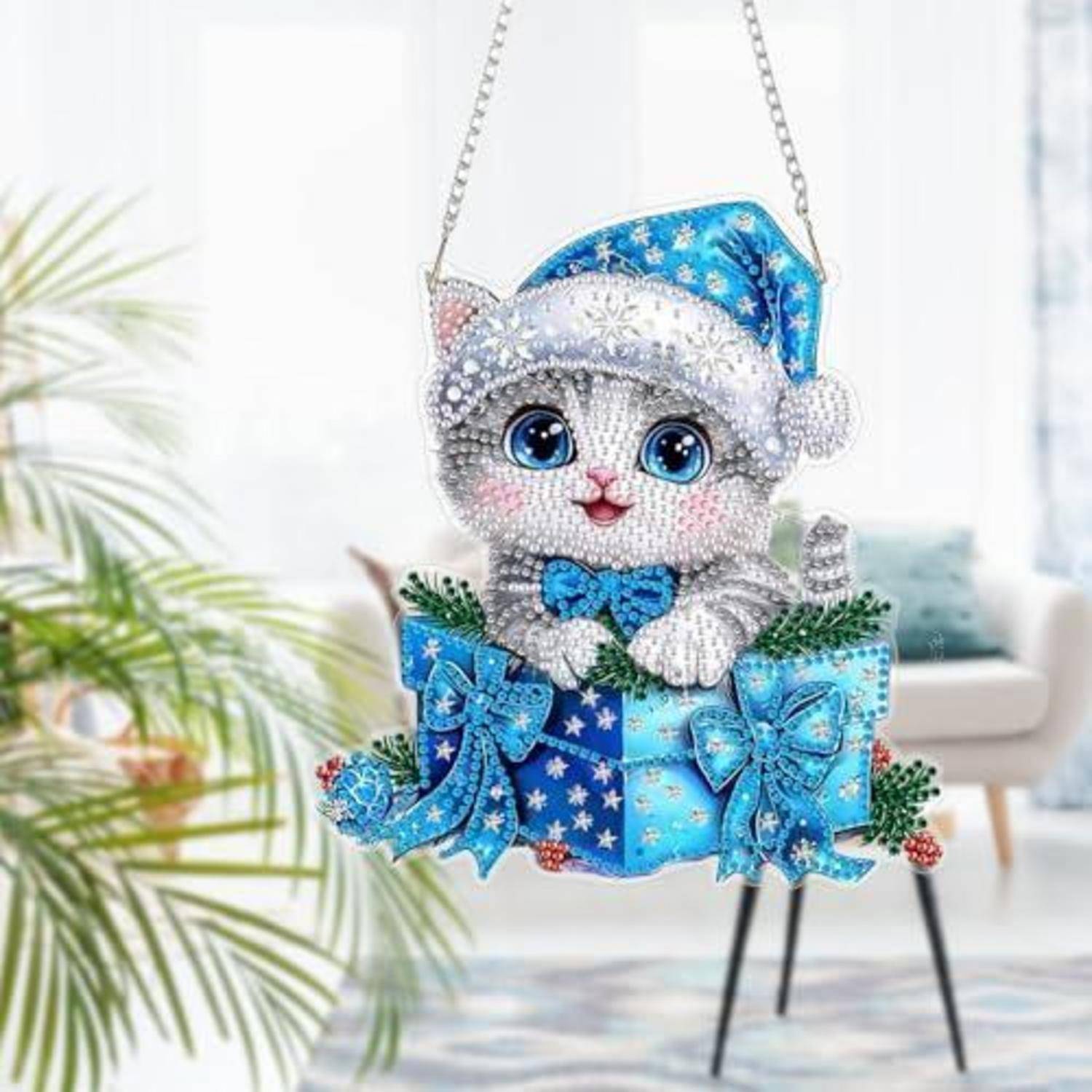 Ein glitzerndes katzenförmiges Weihnachtsornament mit blauem Weihnachtsmütze und Schleife sitzt in einer blauen Geschenkbox und hängt an einer Kette in einem hellen Raum.