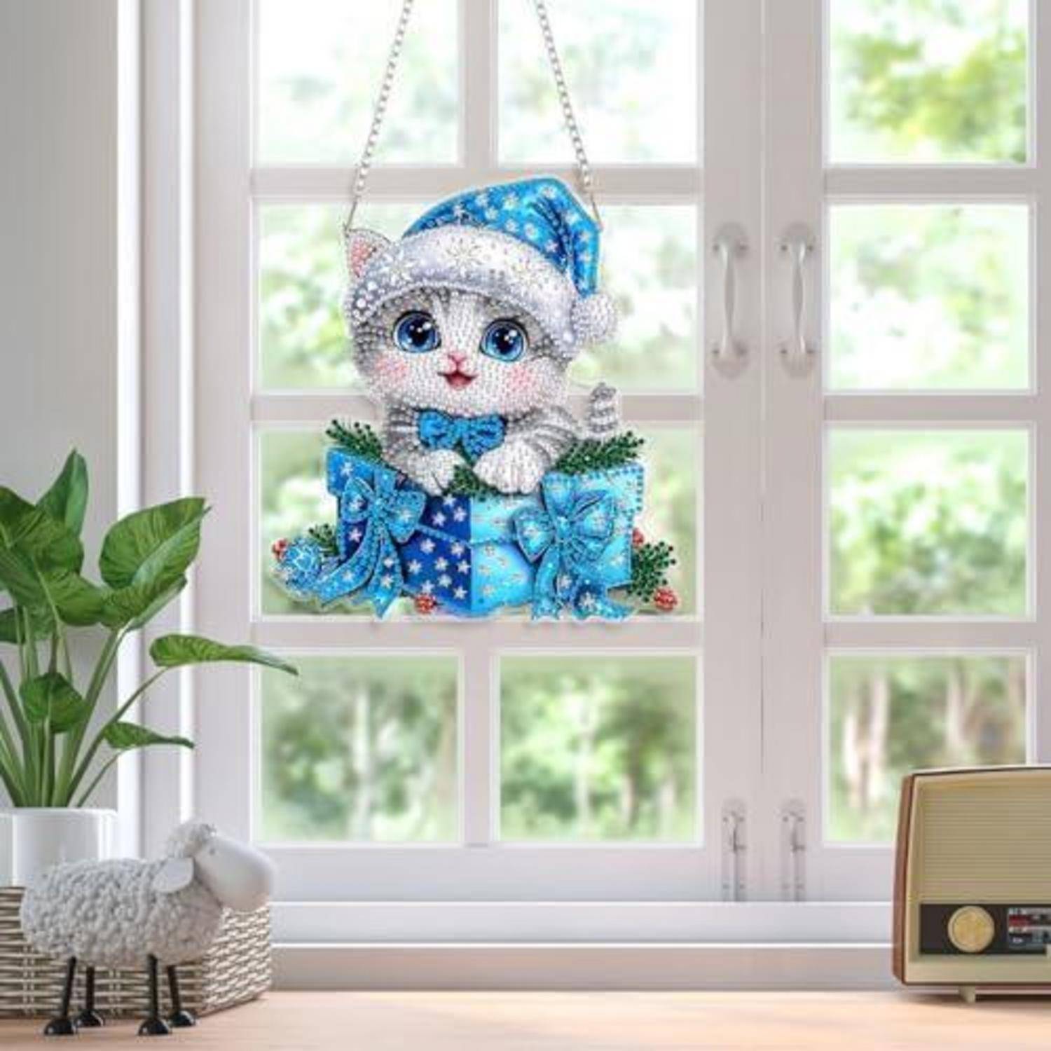 Ein juwelenverziertes, katzenförmiges Weihnachtsornament mit einem blauen Weihnachtsmannhut hängt vor einem hellen Fenster, umgeben von einer Pflanze und einem Vintage-Radio.