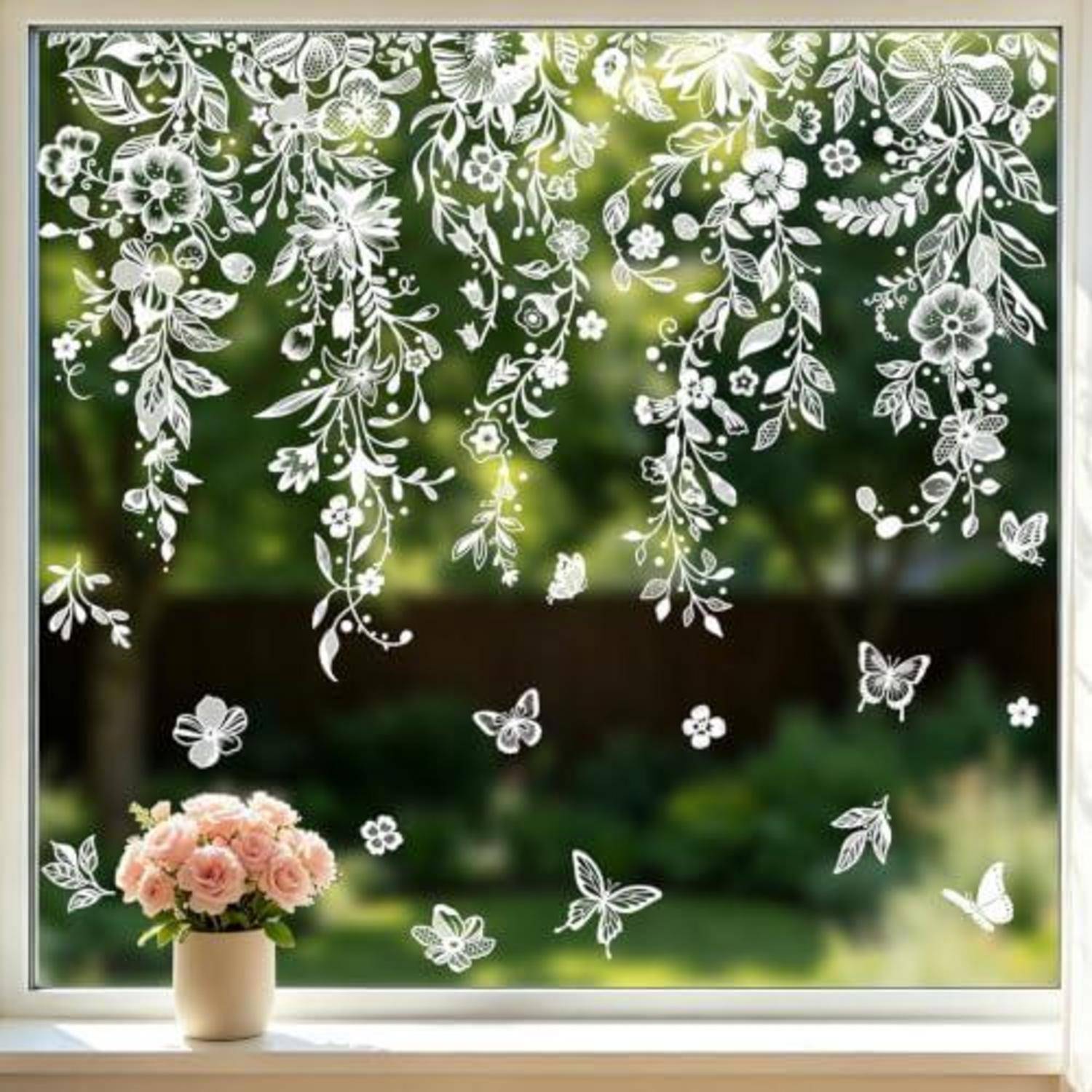 Weißes Blumenwein-Dekoraufkleber auf einem Fenster, das eine kleine Vase mit rosa Rosen rahmt, mit Schmetterlingen und Blumen vor einem unscharfen grünen Außenblick.