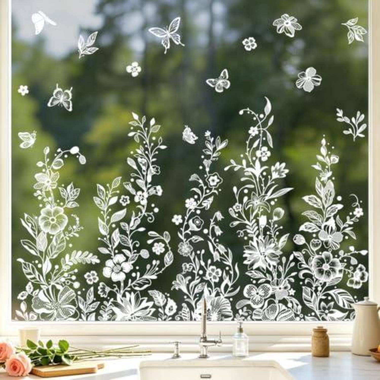 Weißes florales und Schmetterlings-Fensterdekorationspapier über einem Küchenbecken, mit Pflanzen und Blumen entlang des unteren Glases vor einem unscharfen grünen Außenblick.