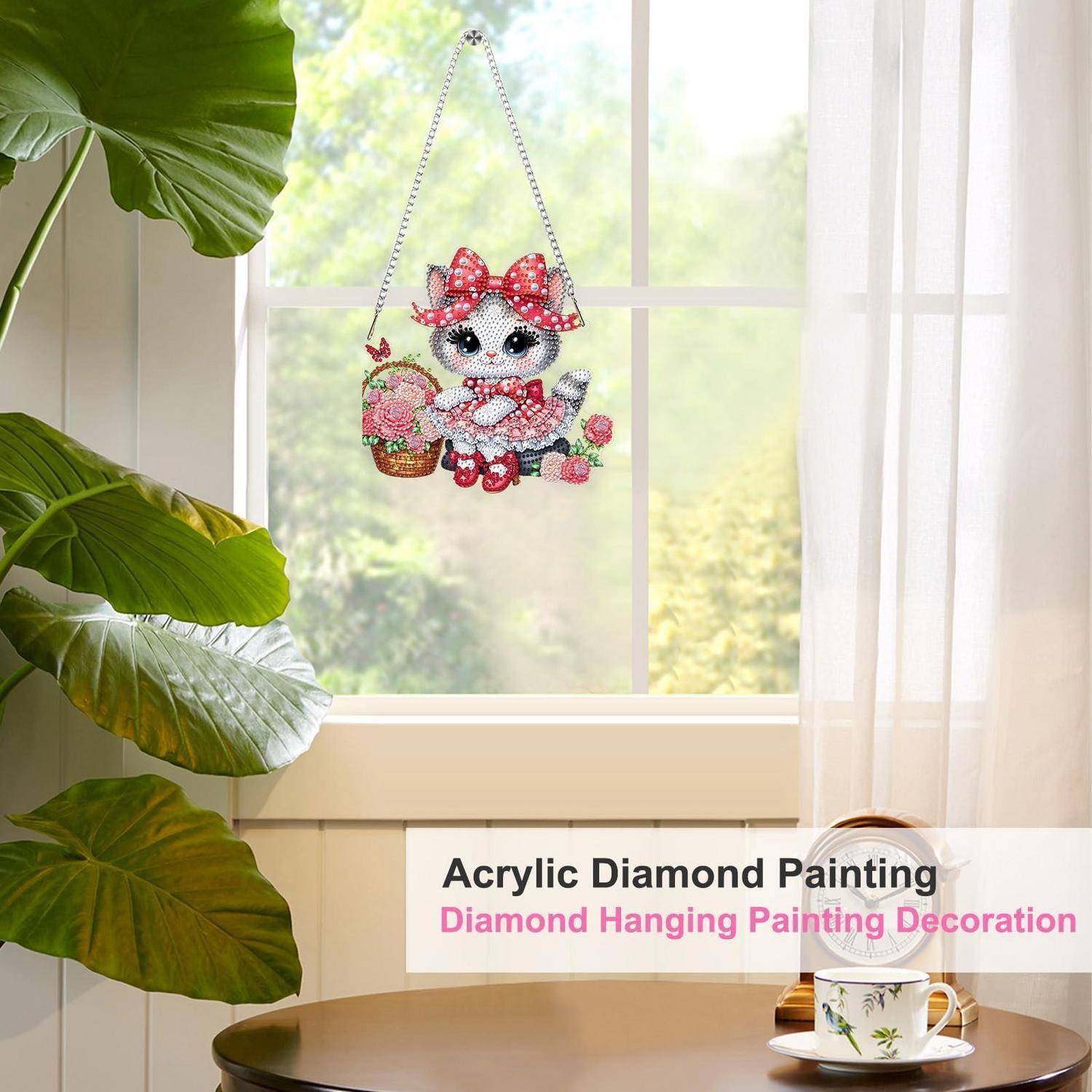 Eine Diamantmalerei-Wanddekoration einer Cartoon-Katze mit einem Korb und einer Schleife, ausgestellt in einer hellen Fensterszene mit einem Tisch und einer Pflanze.