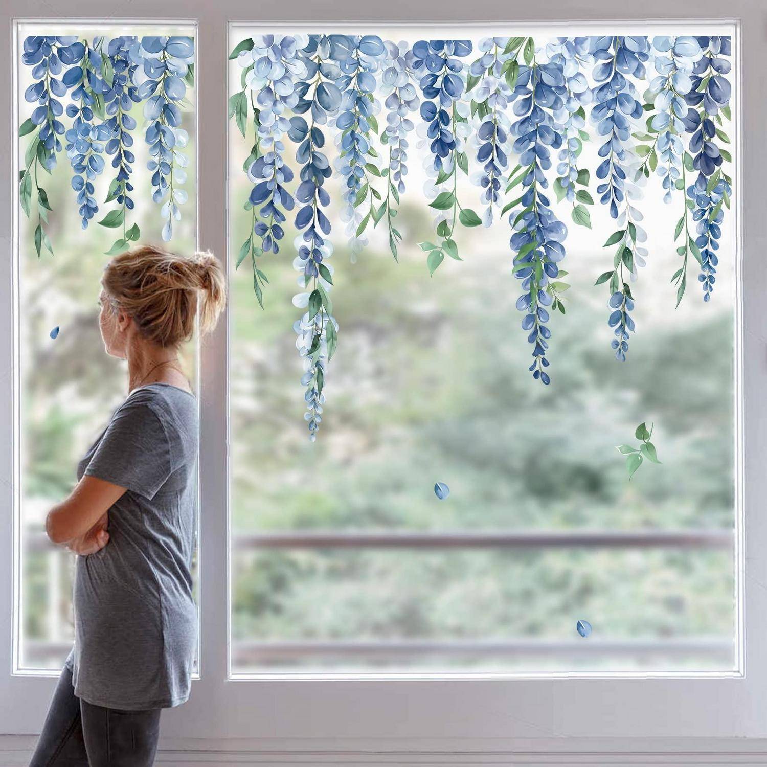 Eine Frau steht am Fenster und blickt nach draußen, mit blauen hängenden Blumendekoration über dem oberen Teil der Glasscheibe.