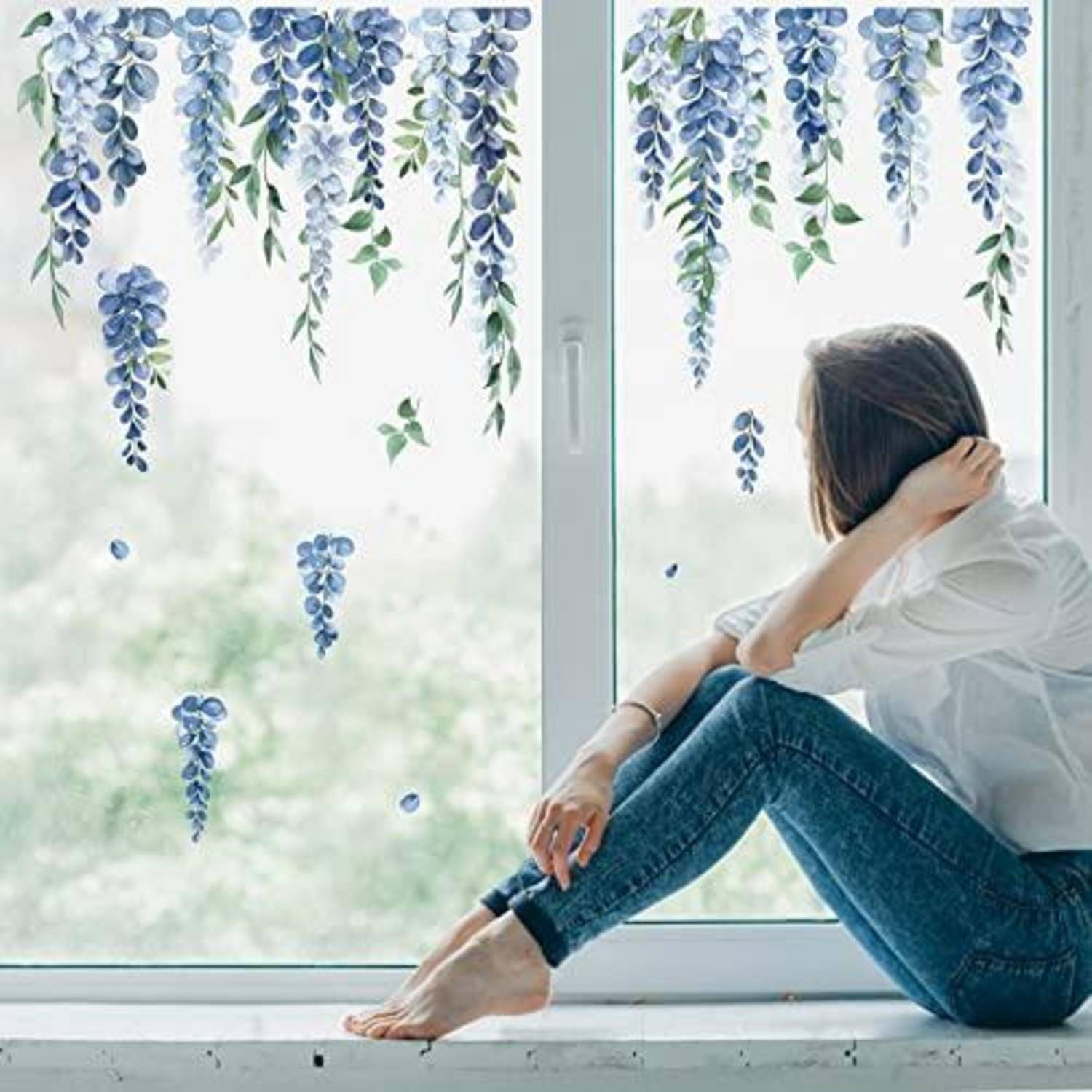 Eine Frau sitzt auf einer Fensterbank und schaut auf dekorative blaue Blumenaufkleber hinaus, die am Fenster hängen, was einen ruhigen, nachdenklichen Moment drinnen andeutet.