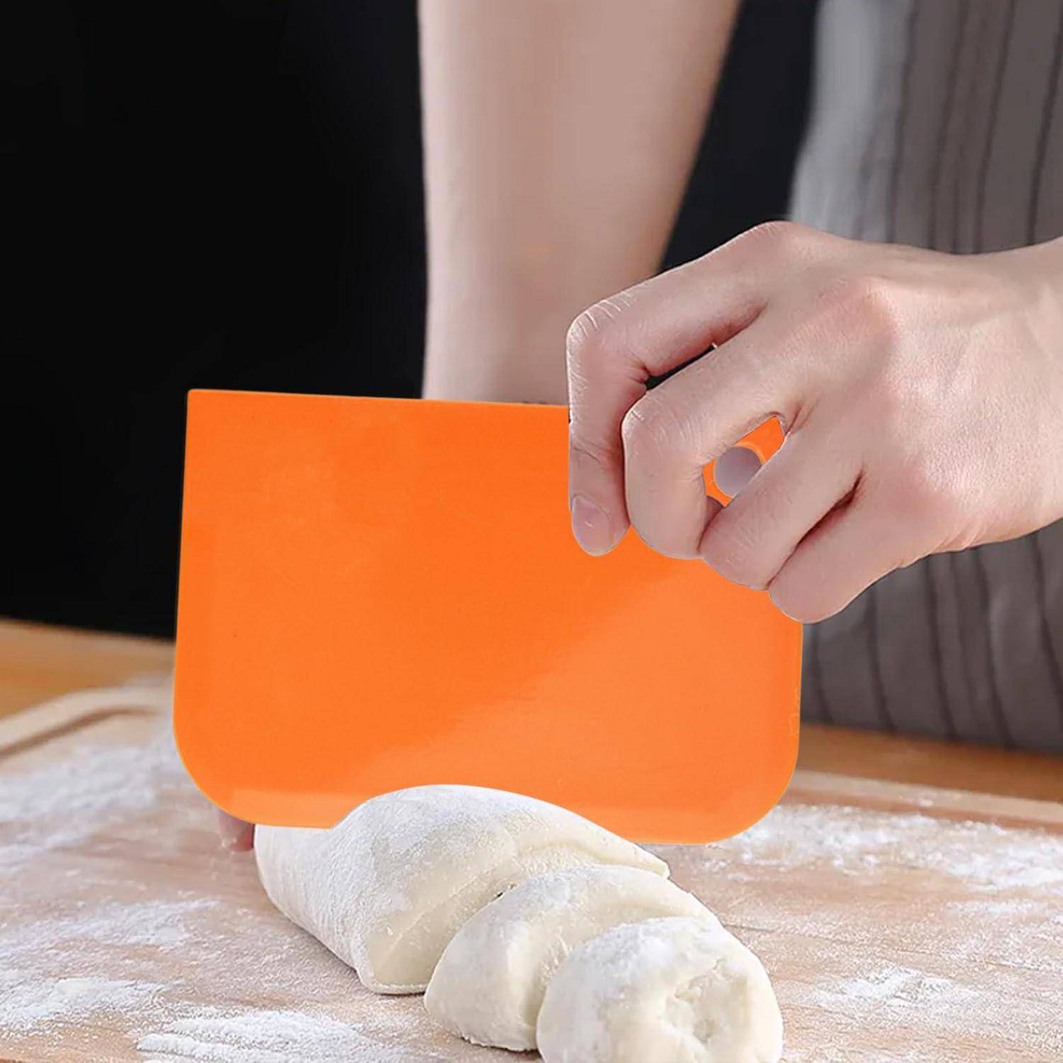 Hände verwenden einen orangefarbenen Teigschaber, um ausgerollten Teig auf einer bemehlten Oberfläche zu schneiden.