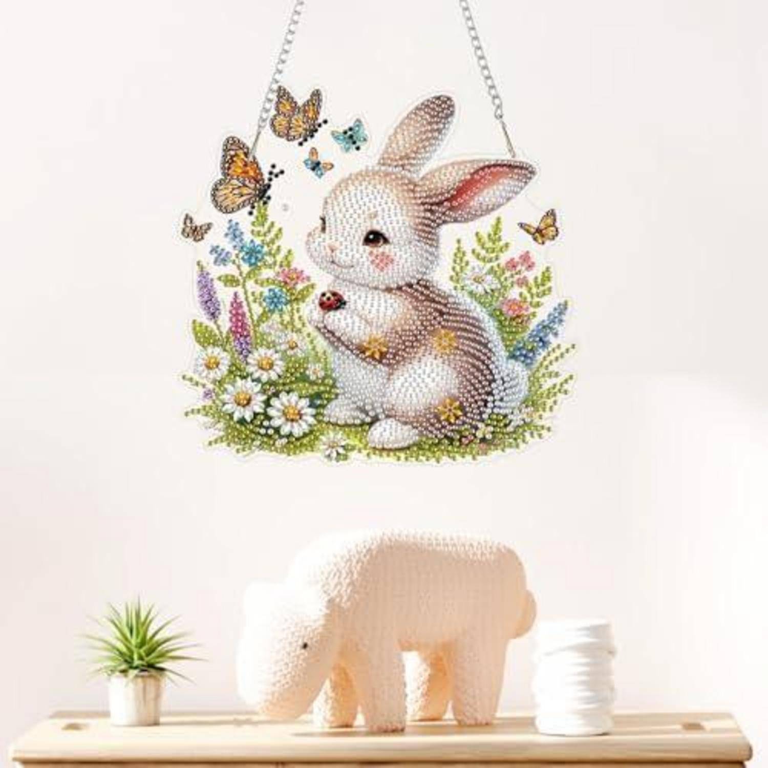 Dekorative Kaninchenkunst mit Schmetterlingen und Blumen, die über einem Regal hängt, mit einer kleinen Topfpflanze, einer weißen Figur und gefalteten Handtüchern darunter.